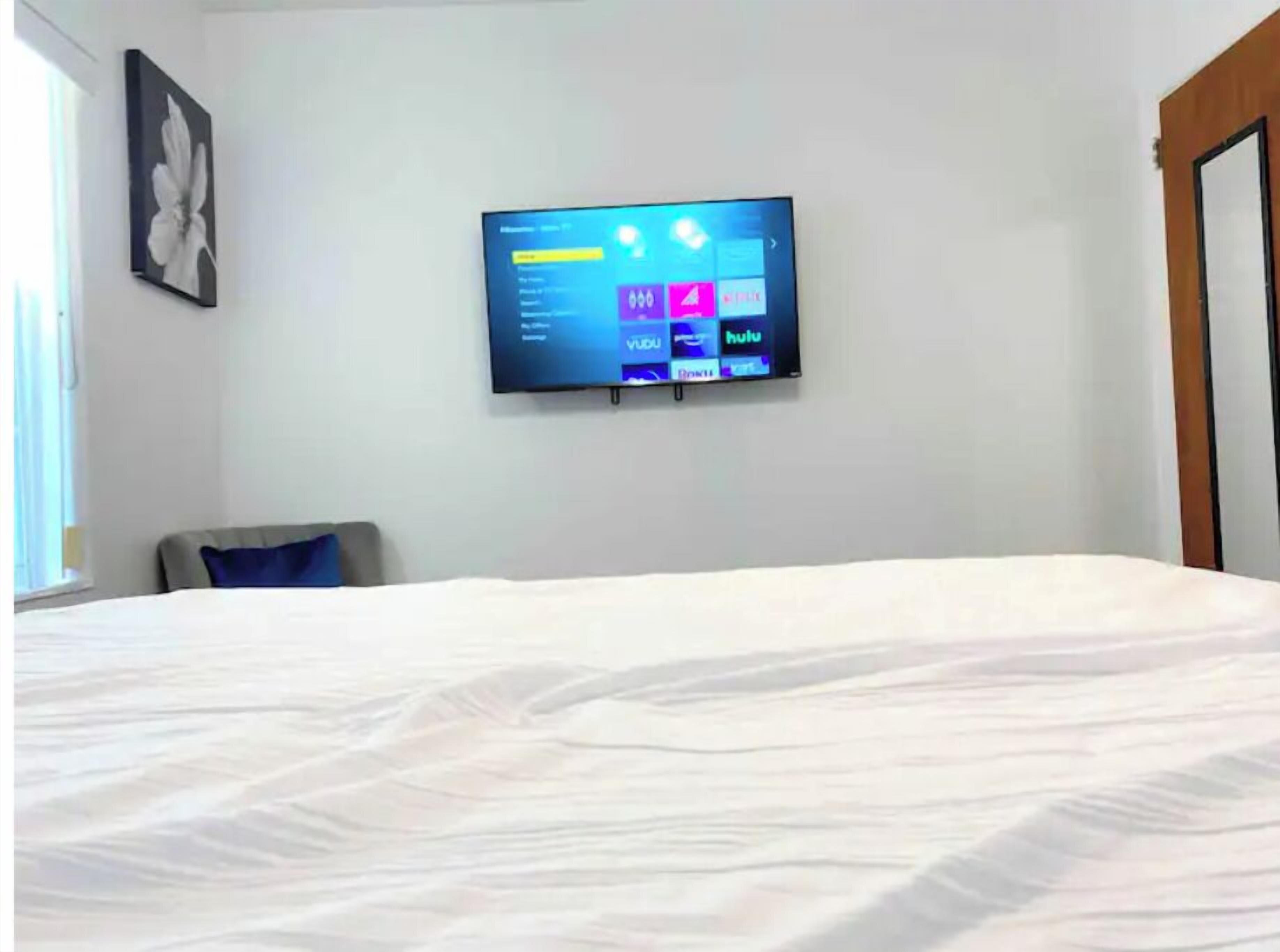 A hotel room with a bed, a flat-screen TV mounted on the wall displaying a TV guide, and a window on the left side.
