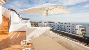 Una terrazza con tavolo e sedie, un ombrellone grande e vista sul mare e sugli edifici.