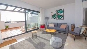 Een moderne woonkamer met een grijze bank, een klein rond tafeltje en een vloerlamp. Er zijn grote ramen en een zichtbaar balkon.