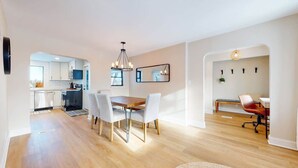A modern kitchen with white cabinets, a dining area with a wooden table and white chairs, and a living area with a sofa and a chair.
