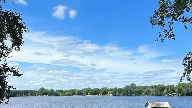 A lakeside seating area with Adirondack chairs and a fire pit, overlooking a lake with a wooden dock and a birdhouse.