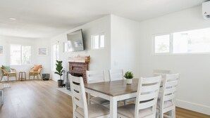 Un espace de repas moderne avec une table en bois et des chaises blanches, une cheminée en briques et un téléviseur fixé au mur.