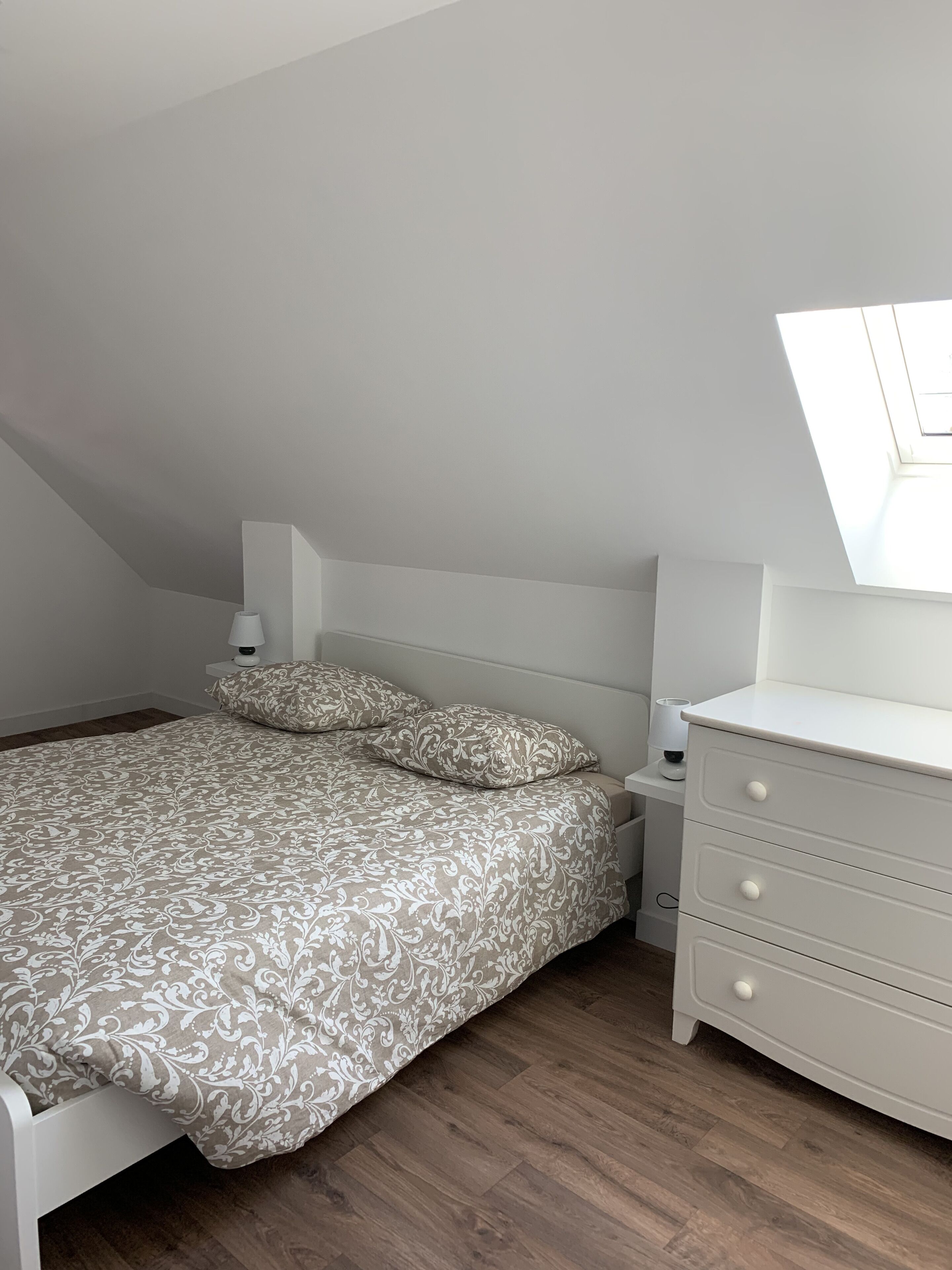 Un dormitorio con techo inclinado, una cama con ropa de cama estampada, una mesita de noche con lámpara y un aparador con cajones.