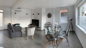 A modern living room with a grey sofa, a glass dining table with patterned chairs, a flat-screen TV, and a large wall clock. - House "LE CH'TI DODO (Guarbecque)