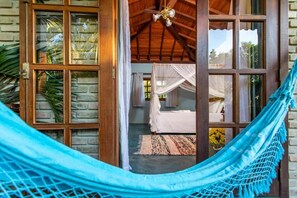 A room with a hammock, a bed with white bedding, and a view of the outside through large windows. - POUSADA SANTO MAR (Porto Seguro)