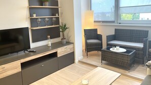 Un salon moderne doté d’un téléviseur à écran plat, d’étagères intégrées ornées d’objets décoratifs, de meubles en osier et d’une grande fenêtre laissant entrer la lumière naturelle.