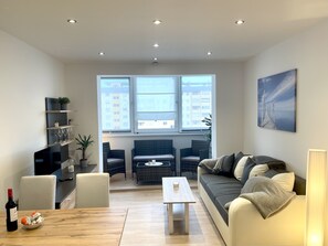 Un moderno salón con sofá, mesa de centro y zona de estar. Hay un ventanal con vistas a edificios, un cuadro en la pared y una botella de vino sobre la mesa.
