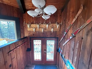 L’intérieur d’un chalet en bois, avec un plafond en bois, un lustre, une porte ornée d’un vitrail et un porte-skis sur le mur de droite.