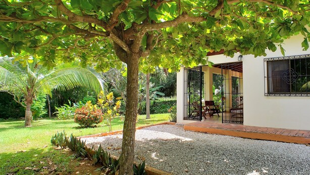 Ein weißes Gebäude mit überdachter Terrasse, umgeben von einem Garten mit verschiedenen Pflanzen und einem Baum.