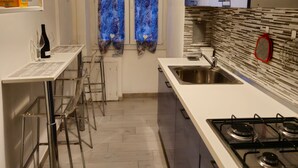 A modern kitchen with a white countertop, stainless steel sink, and gas stove. There are bar stools and a window with blue curtains. - Cozy apartment very close to metro and bus to visit Rome (Roma)