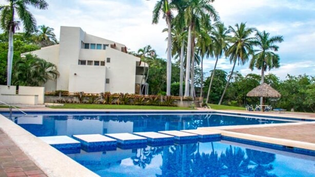 A swimming pool with clear blue water, surrounded by palm trees and a modern building with a flat roof. - Condo Palau by Kivoya (Nuevo Nayarit)