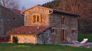 A stone house with a tiled roof, a small porch with two white lounge chairs, and a grassy yard. - Gîte in the heart of the forest, with swimming pool and beautiful views! (VISCOMTAT)