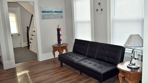 Ein Wohnzimmer mit einer schwarzen Couch, einem kleinen Holztisch, einer Lampe, einem Bild eines Segelboots und einem Leuchtturm-Modell.