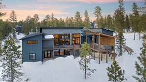 A modern two-story house with a large deck and multiple windows, surrounded by trees and snow.