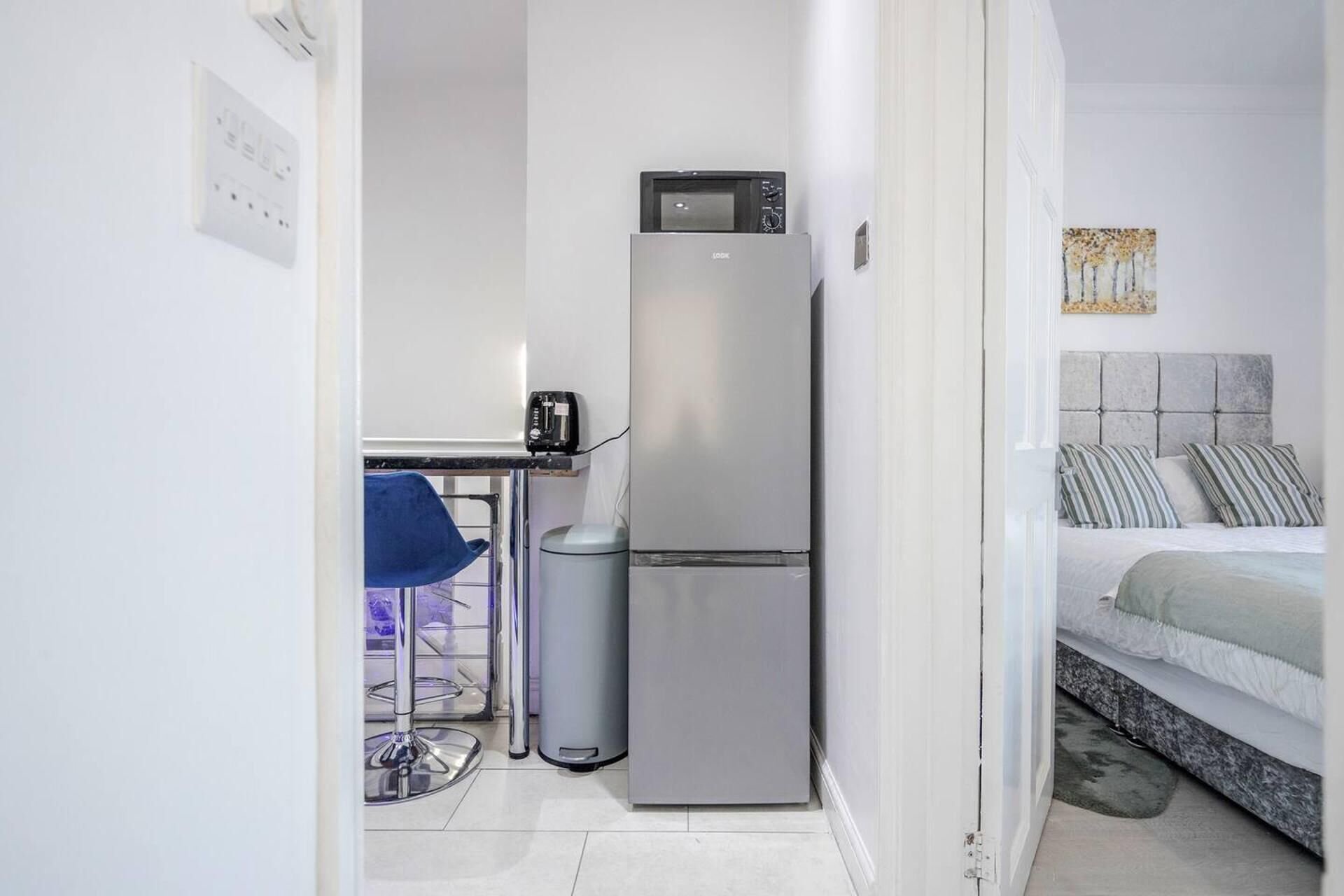 A compact room with a kitchenette, a bed with a grey headboard, and a small seating area with a chair and a trash bin.