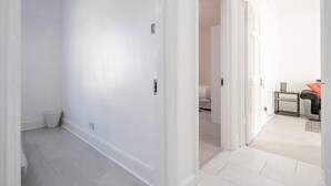 A hallway with white walls and a door leading to a bedroom with a bed and a small table. - Stunning Top 2 Bed Flat Tilbury Central Location (Tilbury)