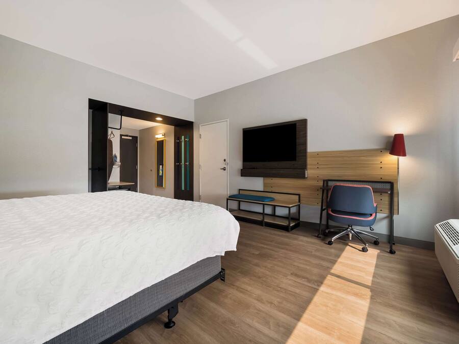 Una habitació d'hotel amb un llit gran, una taula de treball amb una cadira, un televisor pla penjat a la paret i una finestra amb una làmpada vermella a prop.