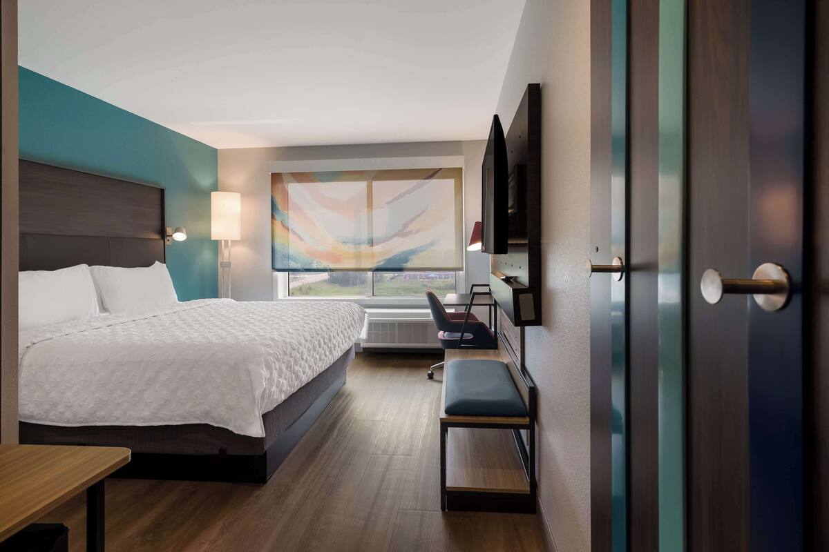 A modern hotel room with a large bed, a desk with a chair, a flat-screen TV, and a large window with a view.