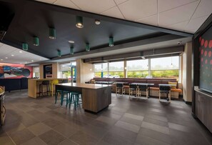 A modern restaurant with a central island, seating area, and large windows offering a view of the outdoors. - Tru By Hilton Novi Detroit (Novi)