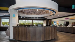 A modern hotel reception area with a curved wooden counter, large windows, and a visible 'Hilton' sign. - Tru By Hilton Novi Detroit (Novi)