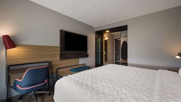 A hotel room with a large bed, a desk with a chair, a television mounted on the wall, and a red lamp.
