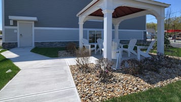 A covered patio area with white chairs and a table, surrounded by a gravel and grass garden.