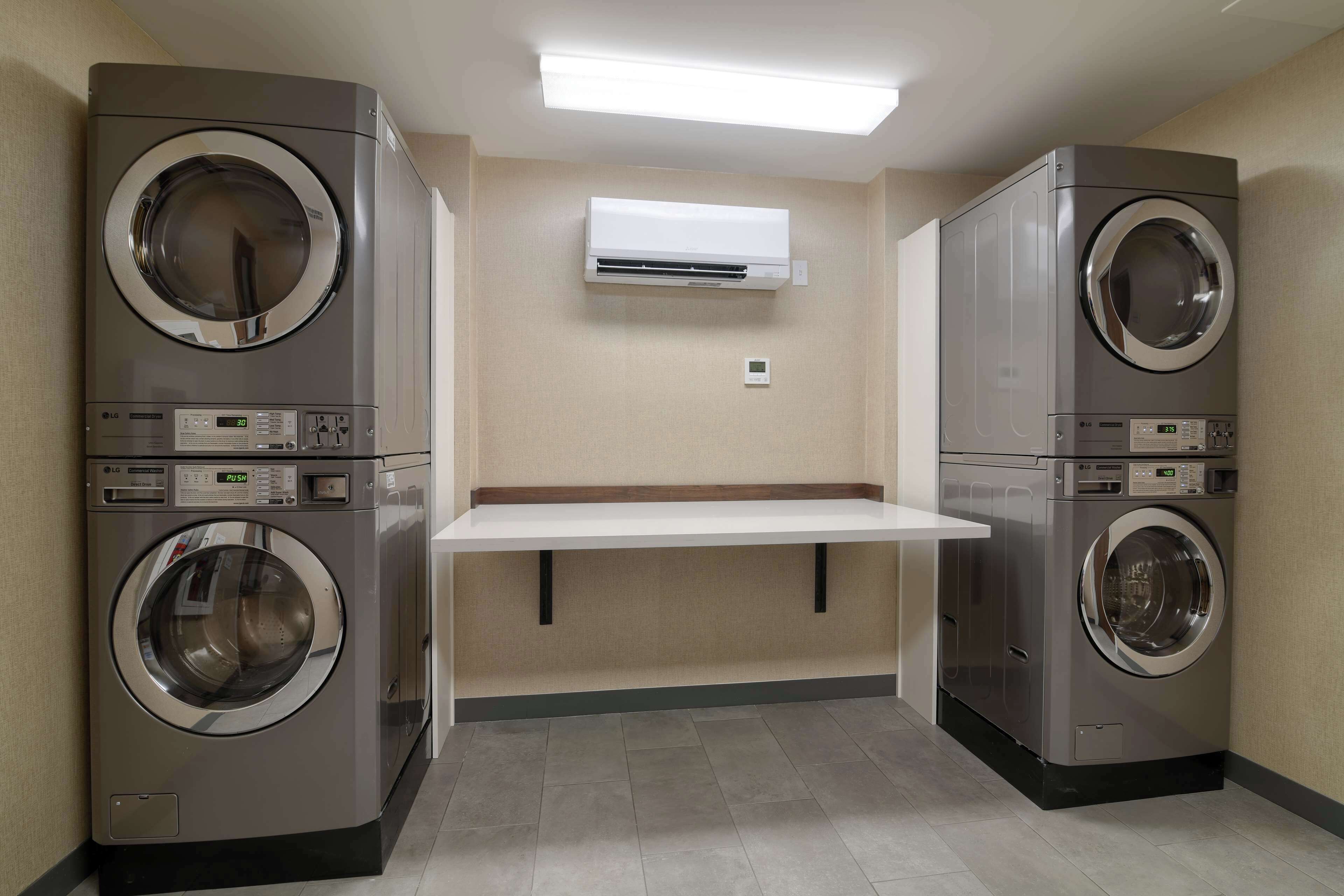 A laundry room with two stacked front-loading washing machines, a white shelf, and a wall-mounted air conditioner.