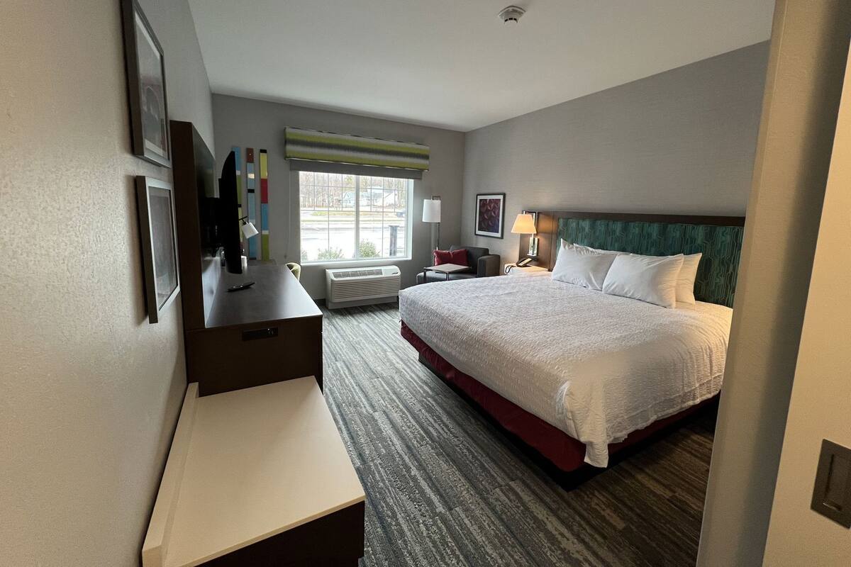 A hotel room with a bed, desk, chair, bedside table, lamp, and a window.