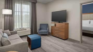 A hotel room with a grey sofa, a blue ottoman, a wooden dresser, a flat-screen TV, and a bed with a blue headboard.