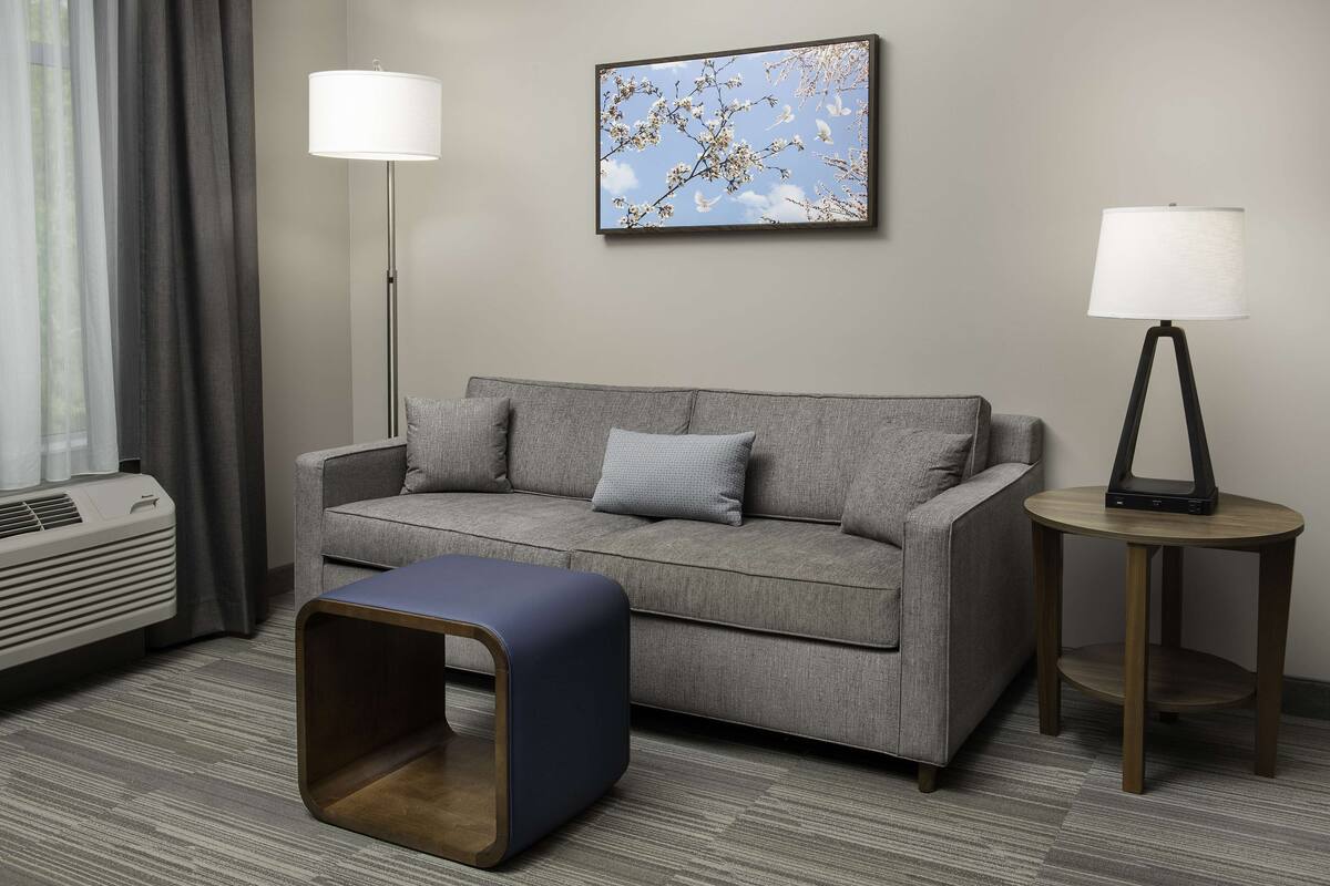 Une chambre d’hôtel moderne avec un canapé gris, deux tables d’appoint avec des lampes, un petit pouf bleu, un téléviseur fixé au mur et une fenêtre avec des rideaux.