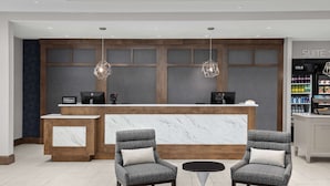 Zona de recepción de hotel con mostrador de mármol, dos sillones tapizados en gris, una mesita redonda y un minibar.