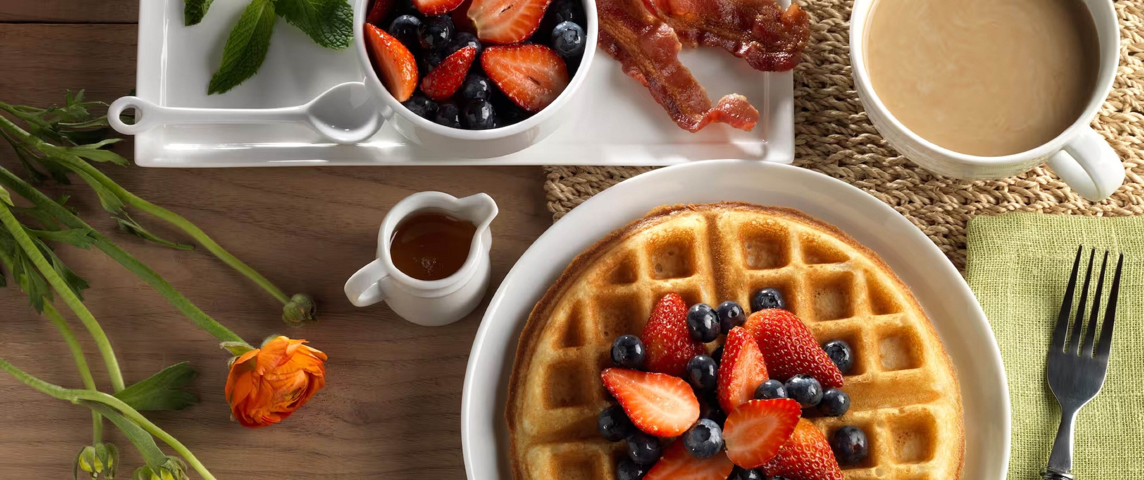A breakfast setting with waffles topped with strawberries and blueberries, a small cup of coffee, and a plate of bacon.