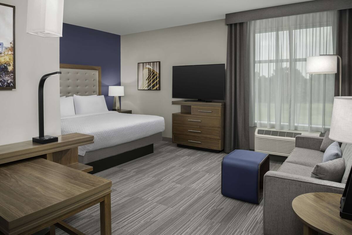Une chambre d’hôtel avec un lit, une télévision, un bureau, un canapé et une fenêtre avec des rideaux.