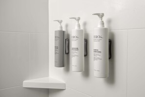 A bathroom with white tiles and three white bottles of shampoo and conditioner on a wall-mounted shelf. - Homewood Suites By Hilton Greenville, NC (Greenville)