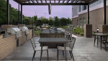 Un patio exterior con una barbacoa de ladrillo, una mesa con cubierta de vidrio y sillas.