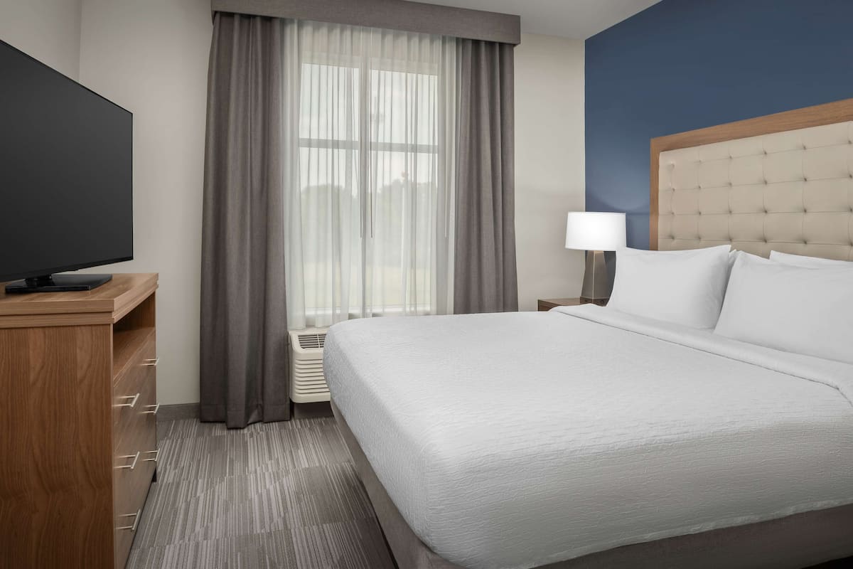 Une chambre d’hôtel dotée d’un grand lit, d’un téléviseur à écran plat, d’un bureau avec des tiroirs, d’une lampe et d’une fenêtre avec des rideaux.