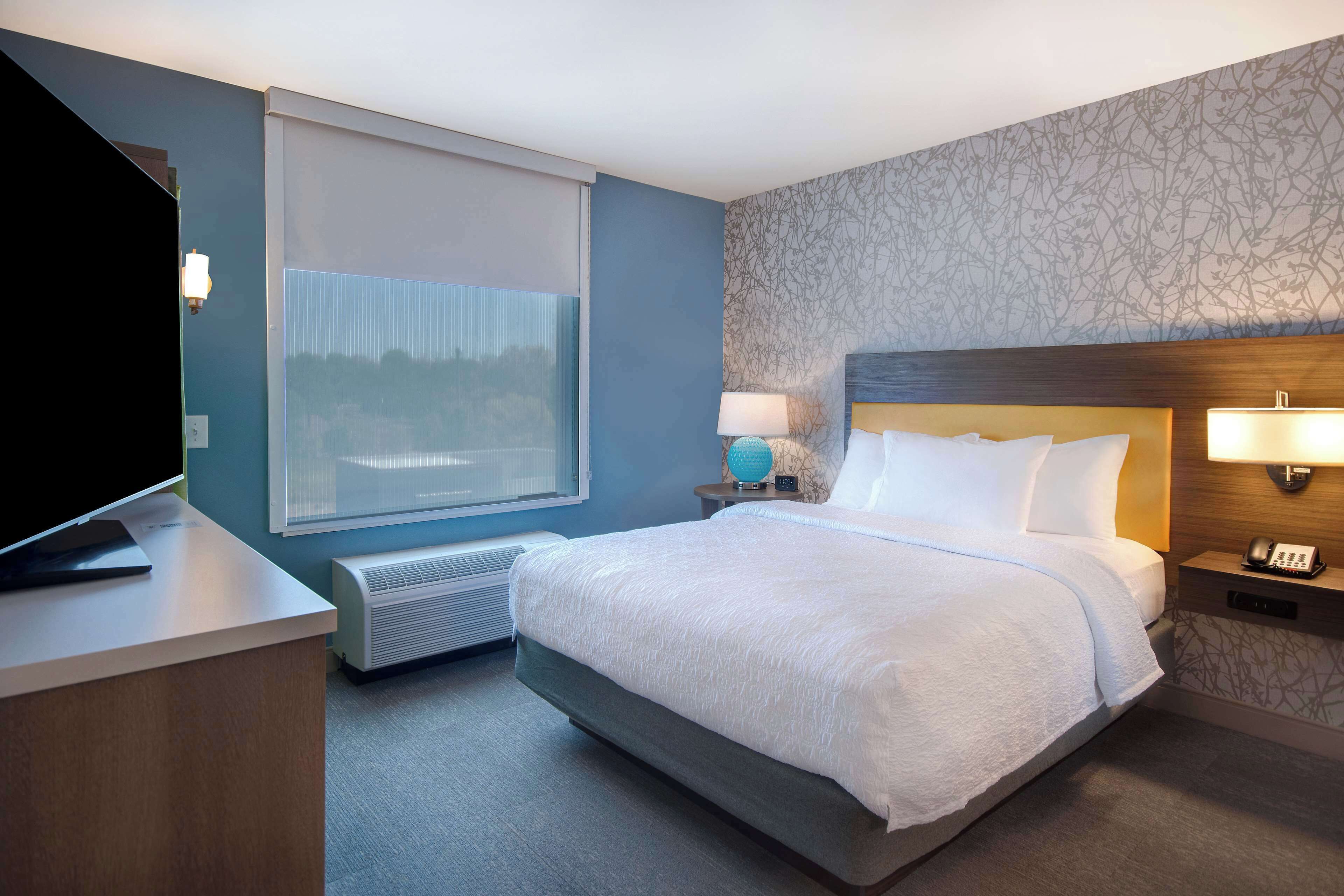 Hotelkamer met een groot bed, een bureau met een flatscreen-tv, een raam met zonwering, een wandlamp en een telefoon op een nachtkastje.