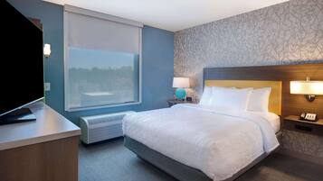 A hotel room with a large bed, a desk with a flat-screen TV, a window with blinds, a wall-mounted lamp, and a phone on a bedside table.