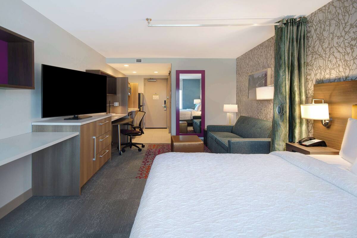 Ein Hotelzimmer mit Bett, Schreibtisch mit Flachbildfernseher, Stuhl, Sofa, Nachttisch mit Lampe und einem durch eine Tür sichtbaren Badezimmer.