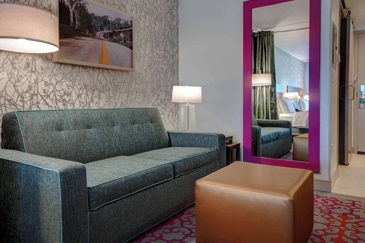 Ein Hotelzimmer mit einem grünen Sofa, einem braunen Hocker, einem großen Spiegel und einem gerahmten Bild an der Wand.