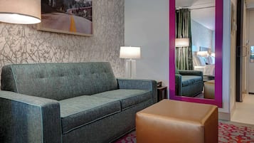Una habitación de hotel con un sofá verde, un otoman marrón, un espejo grande y un cuadro en la pared.