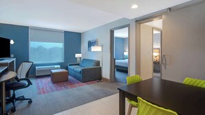 A modern hotel room with a desk, chair, sofa, and a dining table with chairs. - Home2 Suites By Hilton Shepherdsville Louisville South (Louisville)