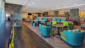 A modern lounge area with a stone accent wall, a long bar, and a seating area with colorful chairs and a sofa. - Home2 Suites By Hilton Shepherdsville Louisville South (Louisville)