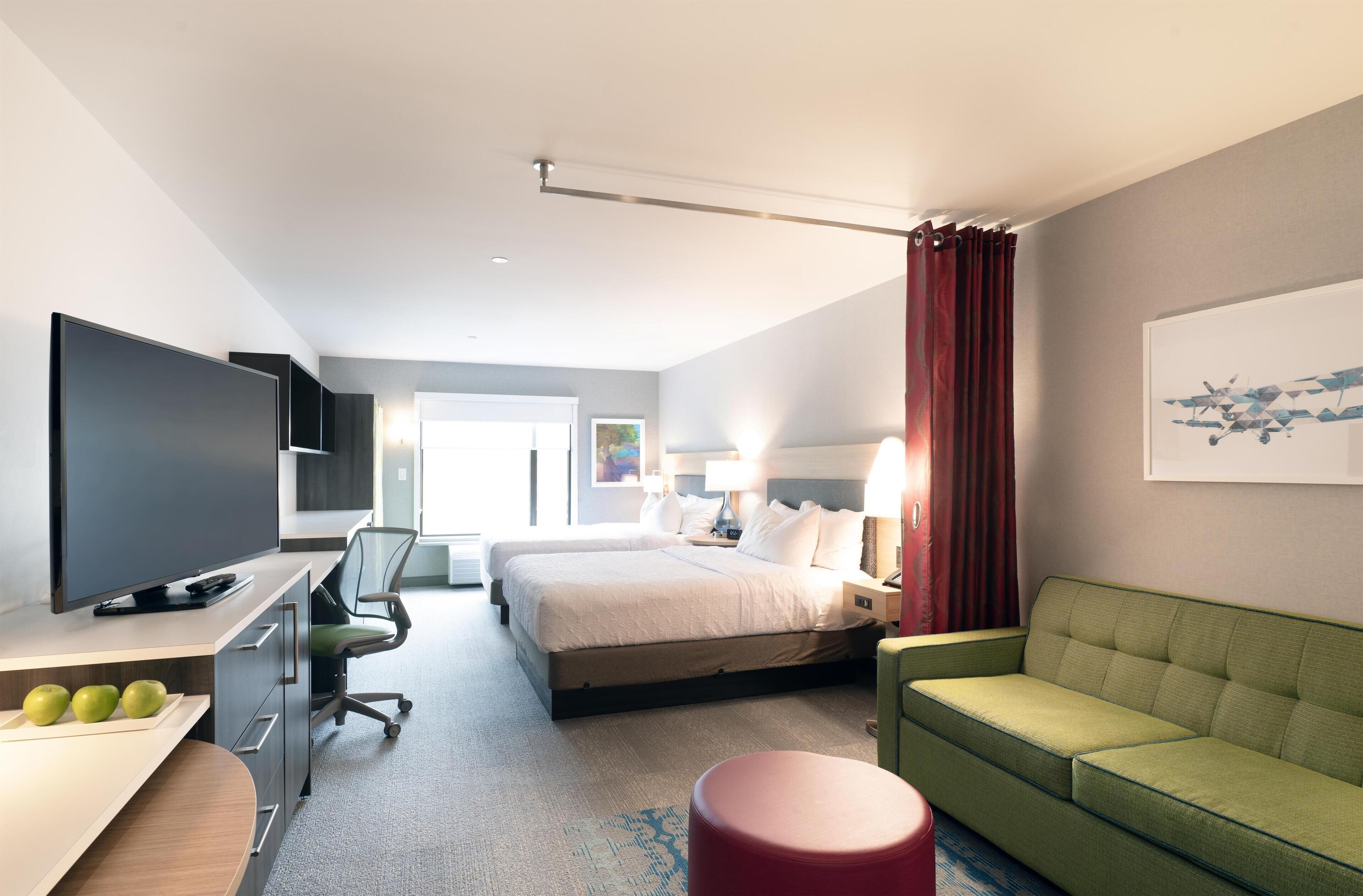 Et hotelværelse med en seng, et skrivebord med tv, en grøn sofa og en rød skammel.