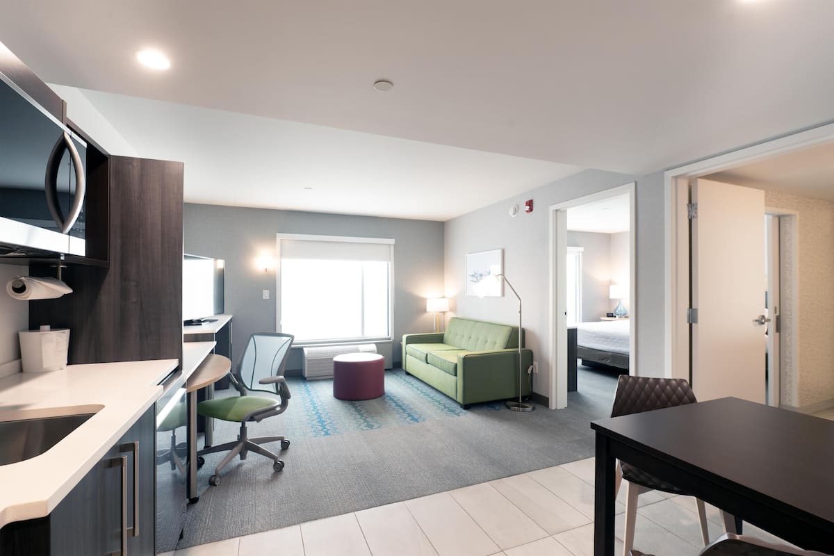 Een moderne hotelkamer met kitchenette, een groene bank, een eettafel en een groot raam dat voor natuurlijk licht zorgt.