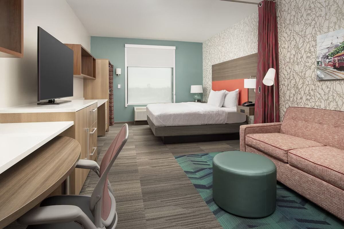 Une chambre d’hôtel comprenant un lit, un bureau avec une télévision, un canapé et un petit pouf.