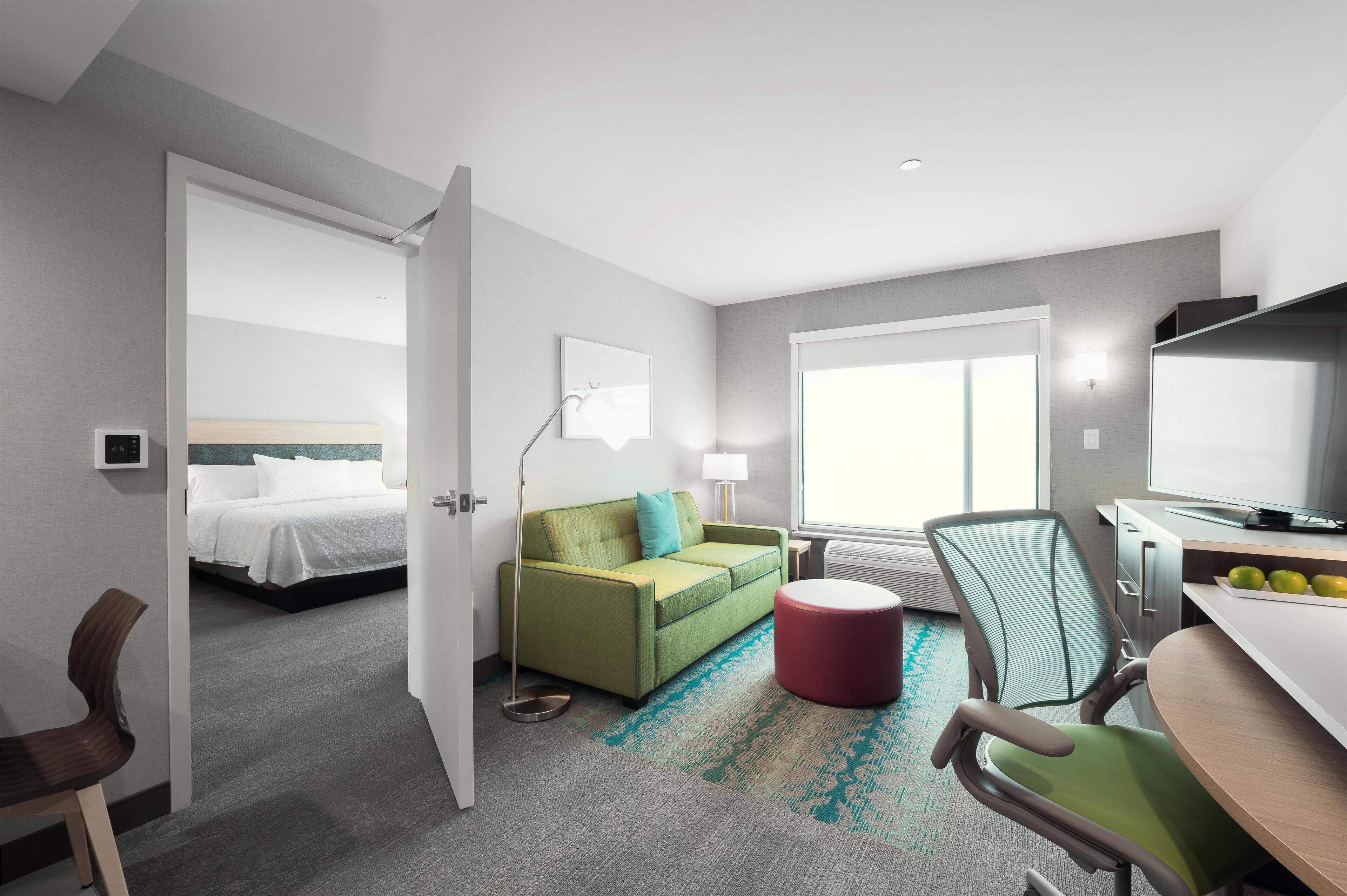 Quarto de hotel com cama, sofá verde, banquinho vermelho, escrivaninha com computador e televisão.