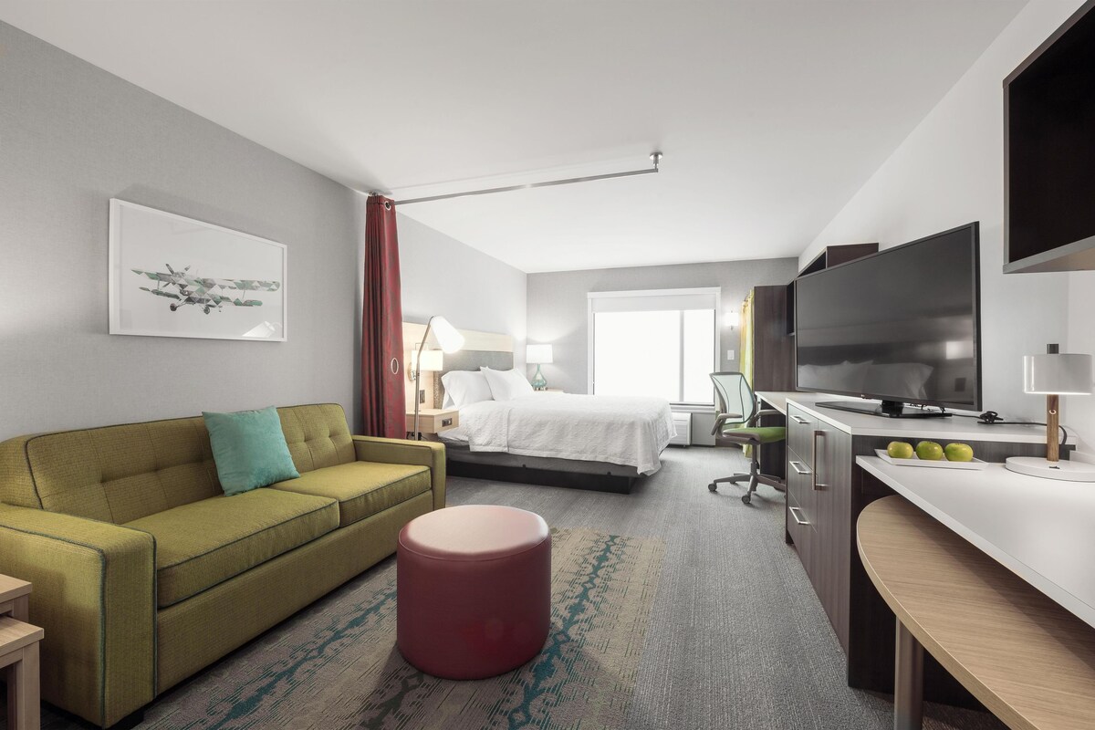 Une chambre d’hôtel avec un lit, un bureau, une télévision, un canapé et un pouf rouge.