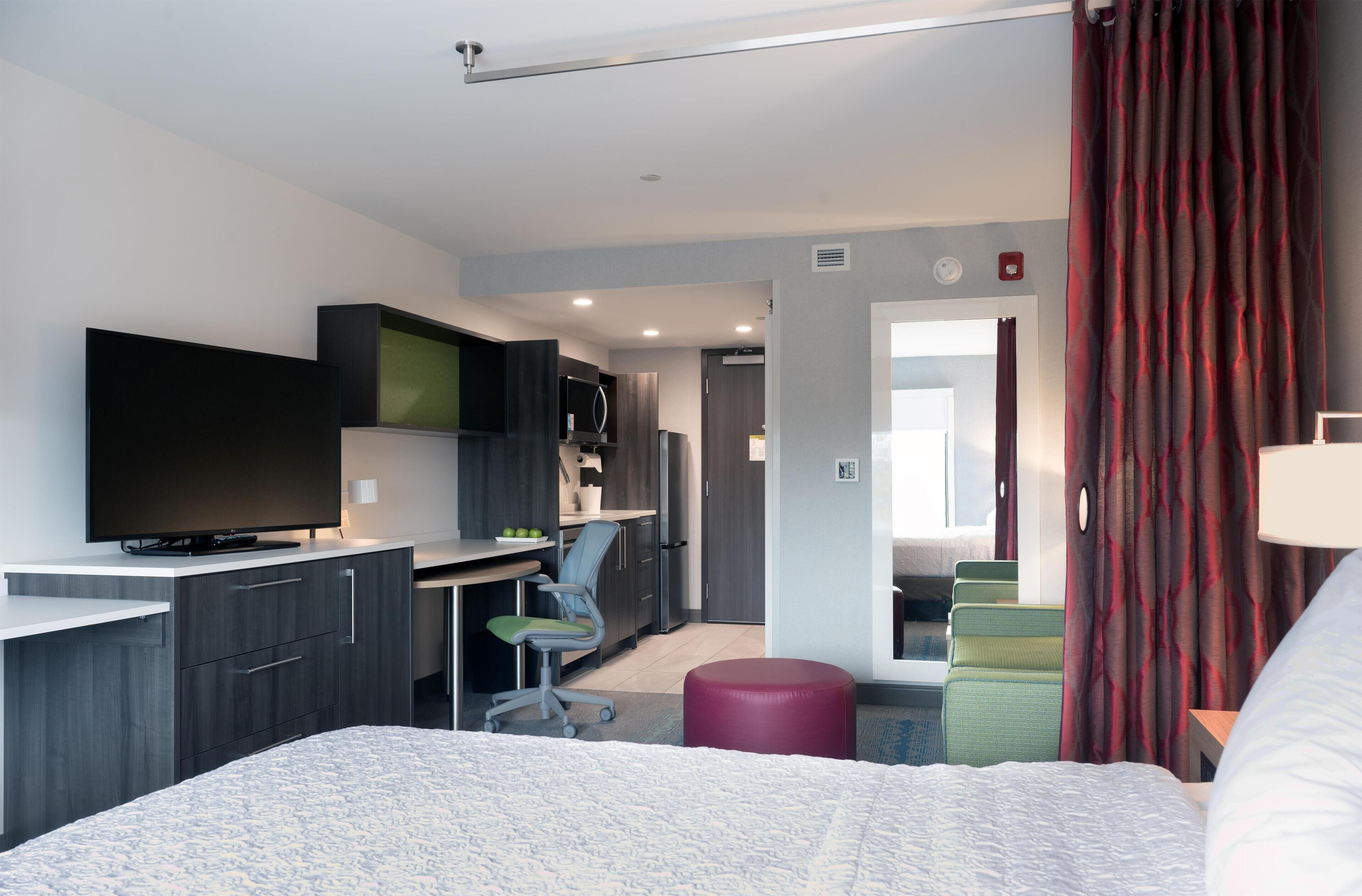 A modern hotel room with a bed, a desk, a chair, a television, a kitchenette, and a bathroom.