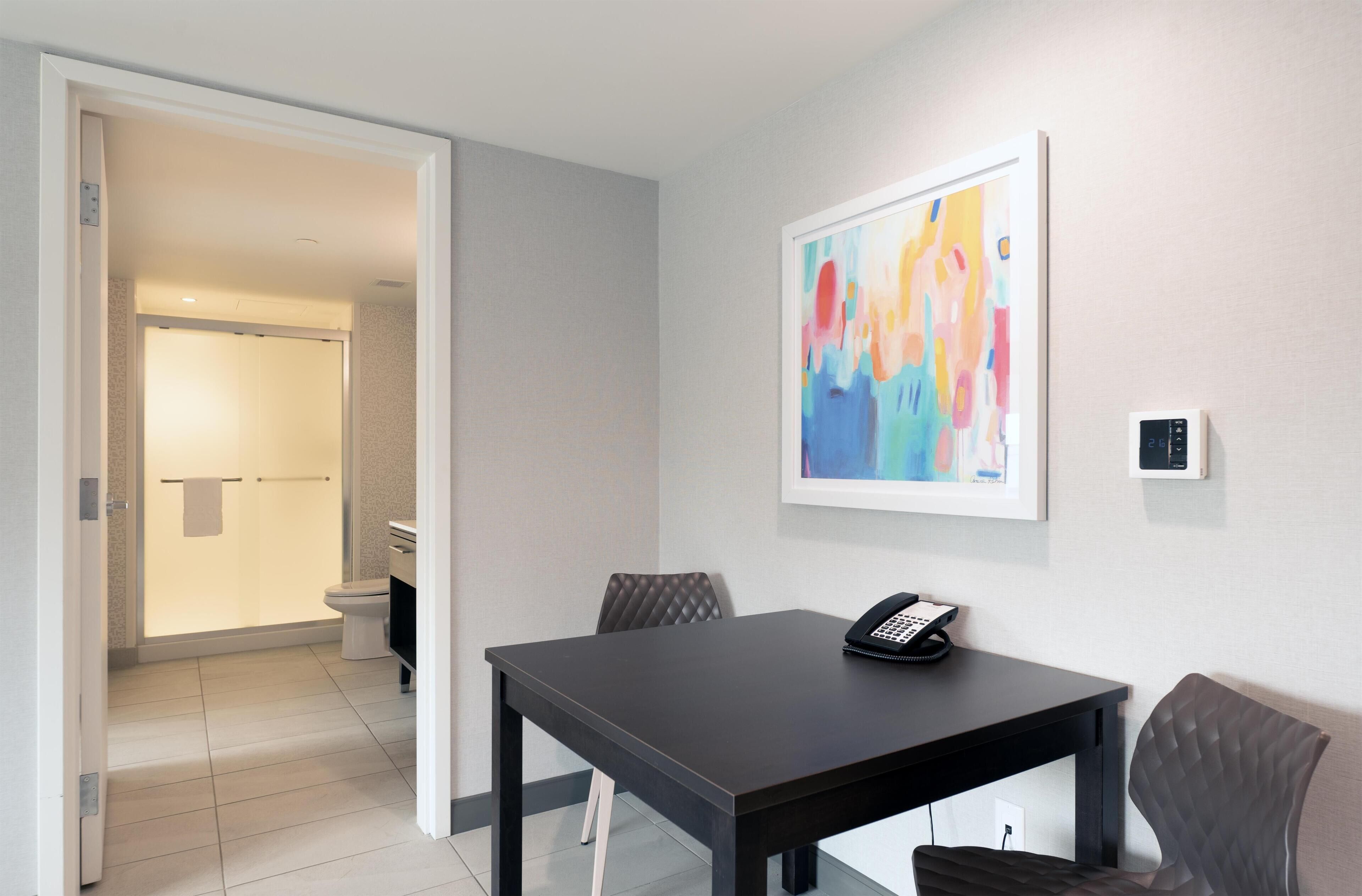A hotel room with a dining table, chairs, a telephone, a painting, and a bathroom visible through an open door.